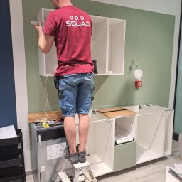 SQUAD S.C. JACEK BARTKOWIAK I KAROL KŁOSEK - Mężczyzna na drabinie montuje białe szafki kuchenne na zielonej ścianie. Widoczne narzędzia i części mebli. Ubrany w czerwoną koszulkę z napisem SQUAD.