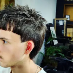 Mężczyzna z awangardową fryzurą: krótka grzywka i dłuższe włosy z tyłu głowy. Widok z profilu, ukazujący precyzyjne cięcie i kształt.