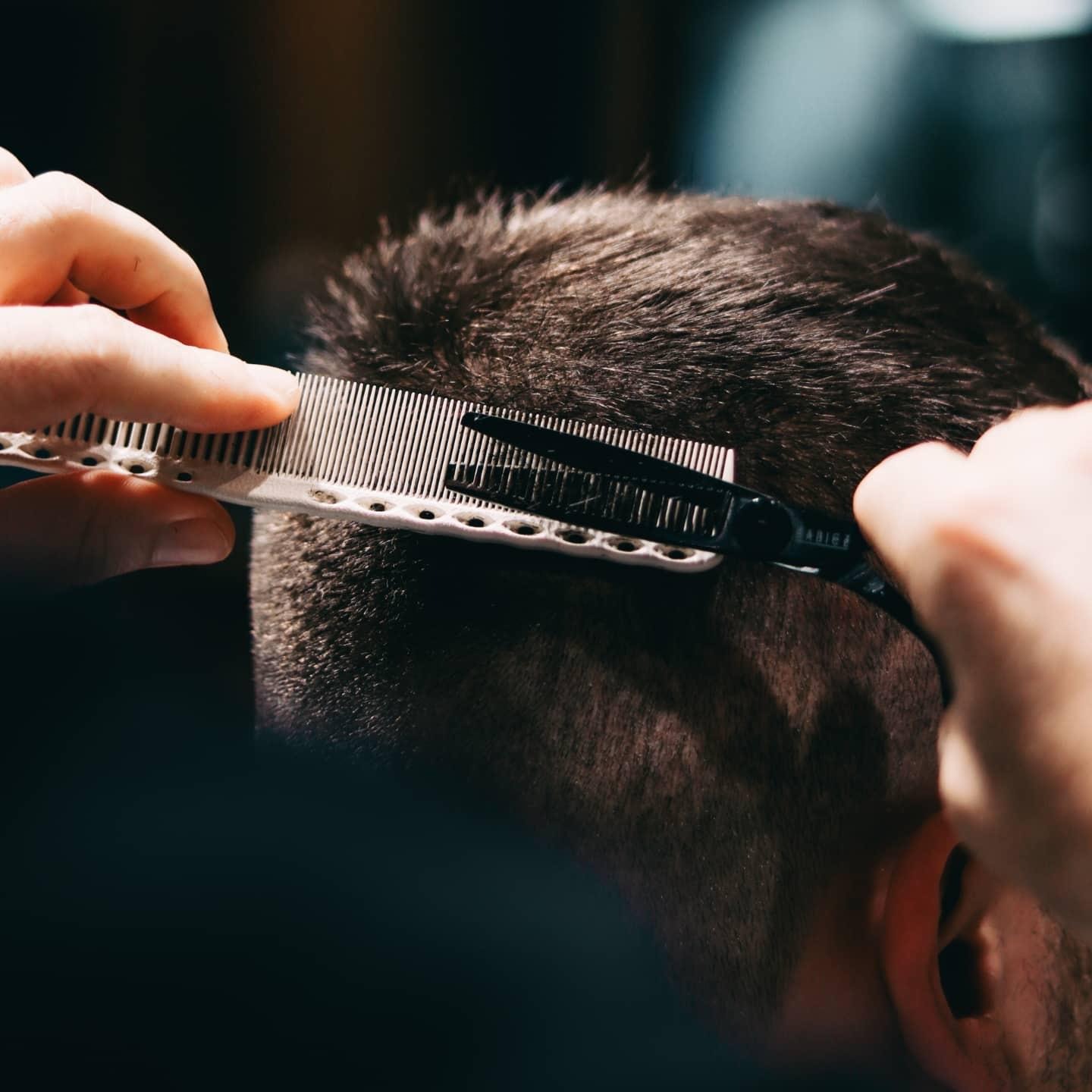 Strzyżenie męskie: dłoń z grzebieniem i nożyczkami modeluje krótkie, ciemne włosy klienta w salonie, ciemne tło, precyzyjne cięcie.