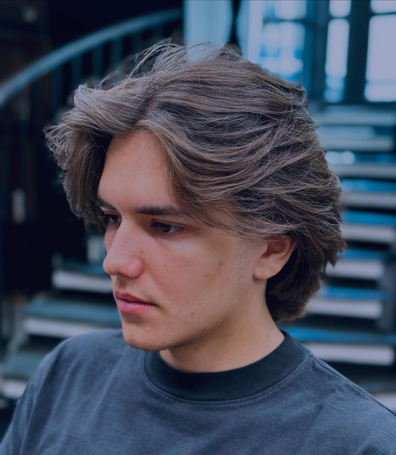 Męska fryzura w stylu lat 90. z brązowymi włosami, widoczna z profilu na tle schodów. Włosy ułożone z wyraźną teksturą i objętością.