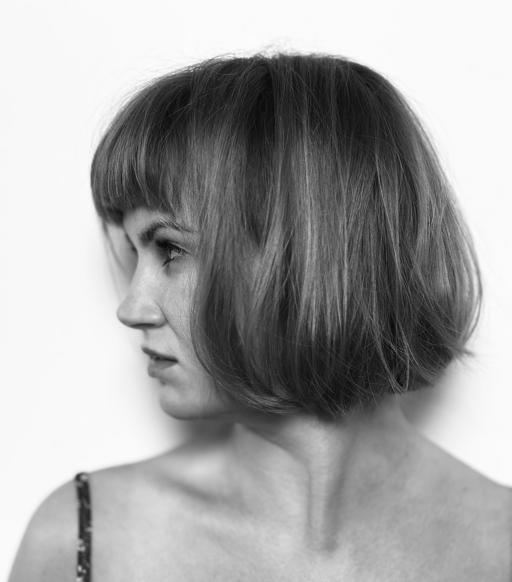 Czarno-białe zdjęcie profilu kobiety z krótką, prostą grzywką i bobem. Delikatny makijaż, subtelny wyraz twarzy, gładka skóra. Minimalistyczne tło.