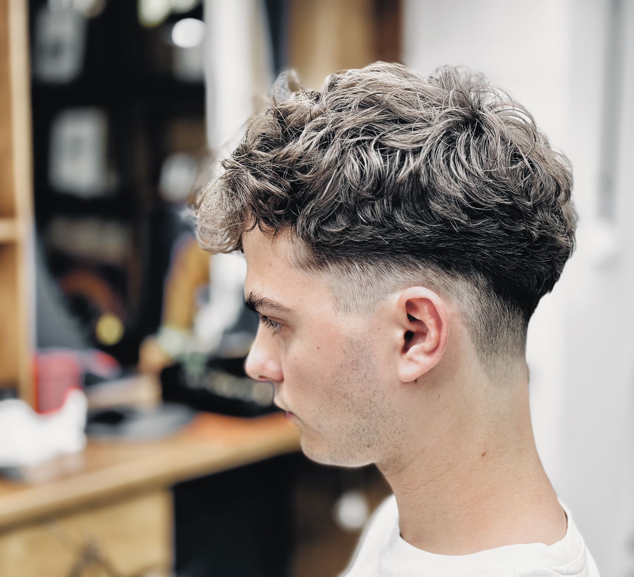 Profil młodego mężczyzny z modną fryzurą typu fade z kręconymi włosami na górze. Włosy w odcieniach szarości i brązu. Twarz gładka, bez zarostu. Tło rozmyte.