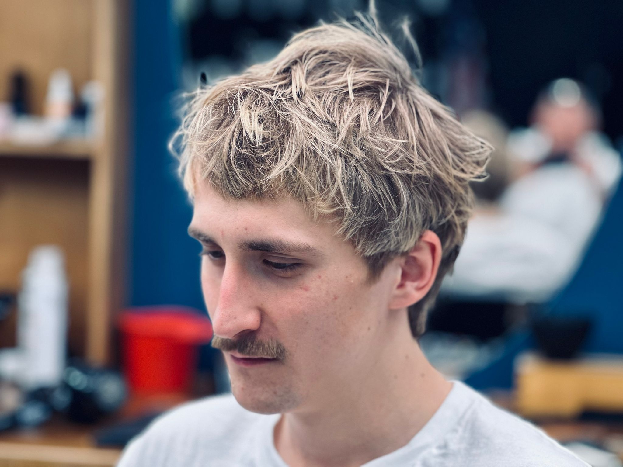 Młody mężczyzna z blond, potarganą fryzurą i wąsami, w białej koszulce, w salonie. Widok z boku, skupienie na włosach i twarzy. W tle akcesoria i meble.