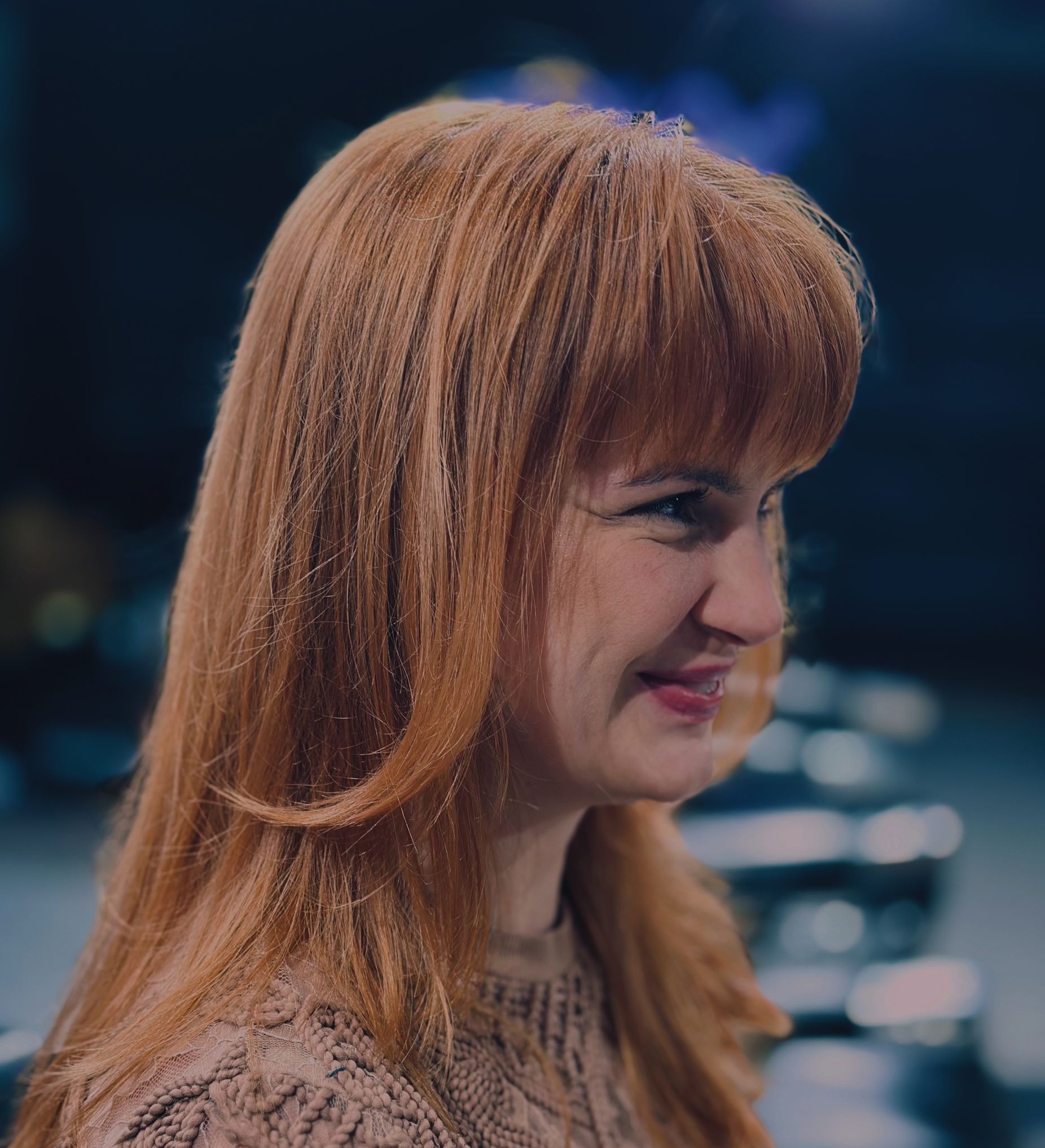 Uśmiechnięta kobieta z rudymi włosami, świeżo po koloryzacji, prezentuje fryzurę z grzywką. Profil, miękkie światło, ciemne tło.