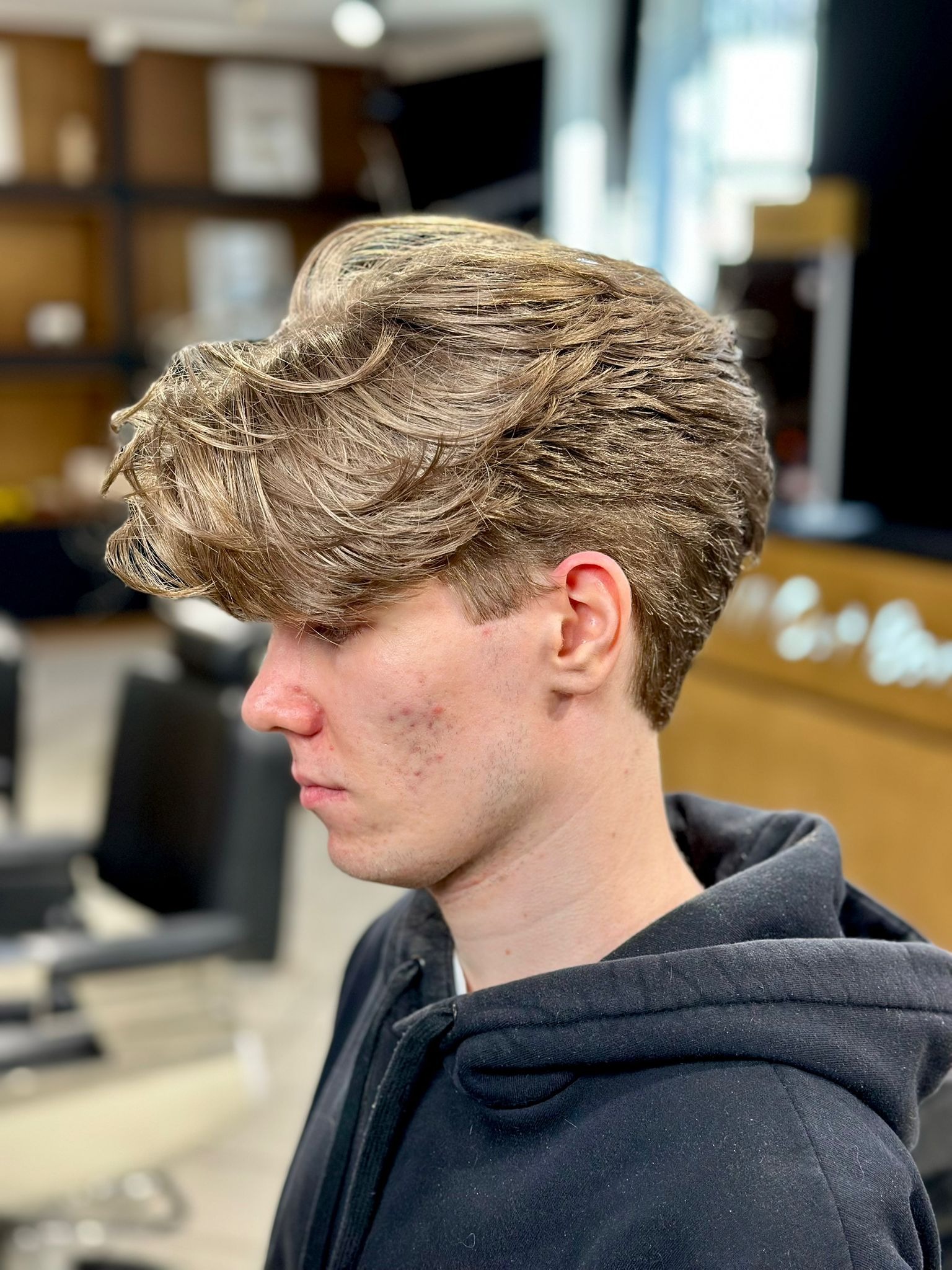 Profil młodego mężczyzny z modną, teksturowaną fryzurą w odcieniach blondu i brązu. W tle widoczne krzesła i elementy wyposażenia salonu.