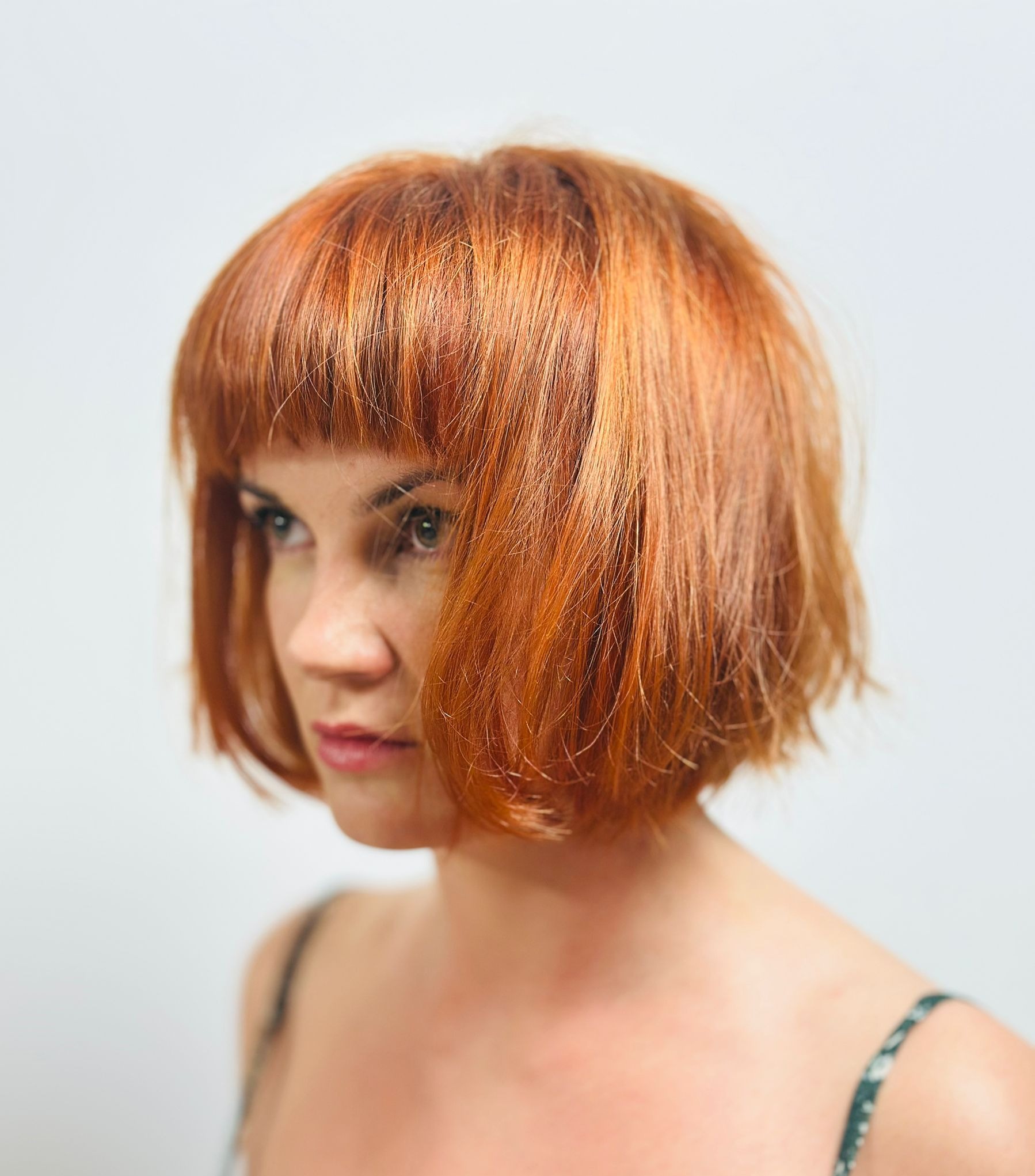 Portret kobiety z krótką, rudą fryzurą z grzywką, skierowanej w lewo, na jasnym tle. Widoczne delikatne cieniowanie włosów i subtelny makijaż.