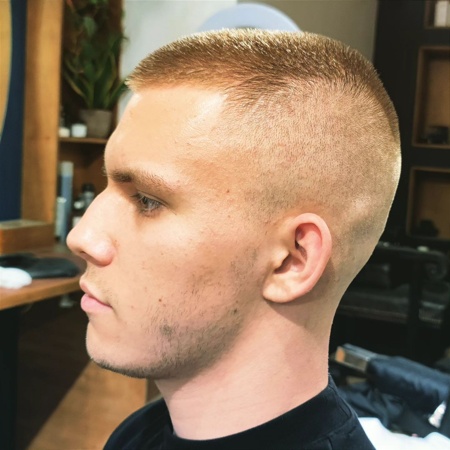 Męska fryzura 'na jeża' z blondu, widok z profilu. Krótkie, równo ostrzyżone włosy, podkreślone naturalnym światłem. W tle widoczne elementy wyposażenia salonu.