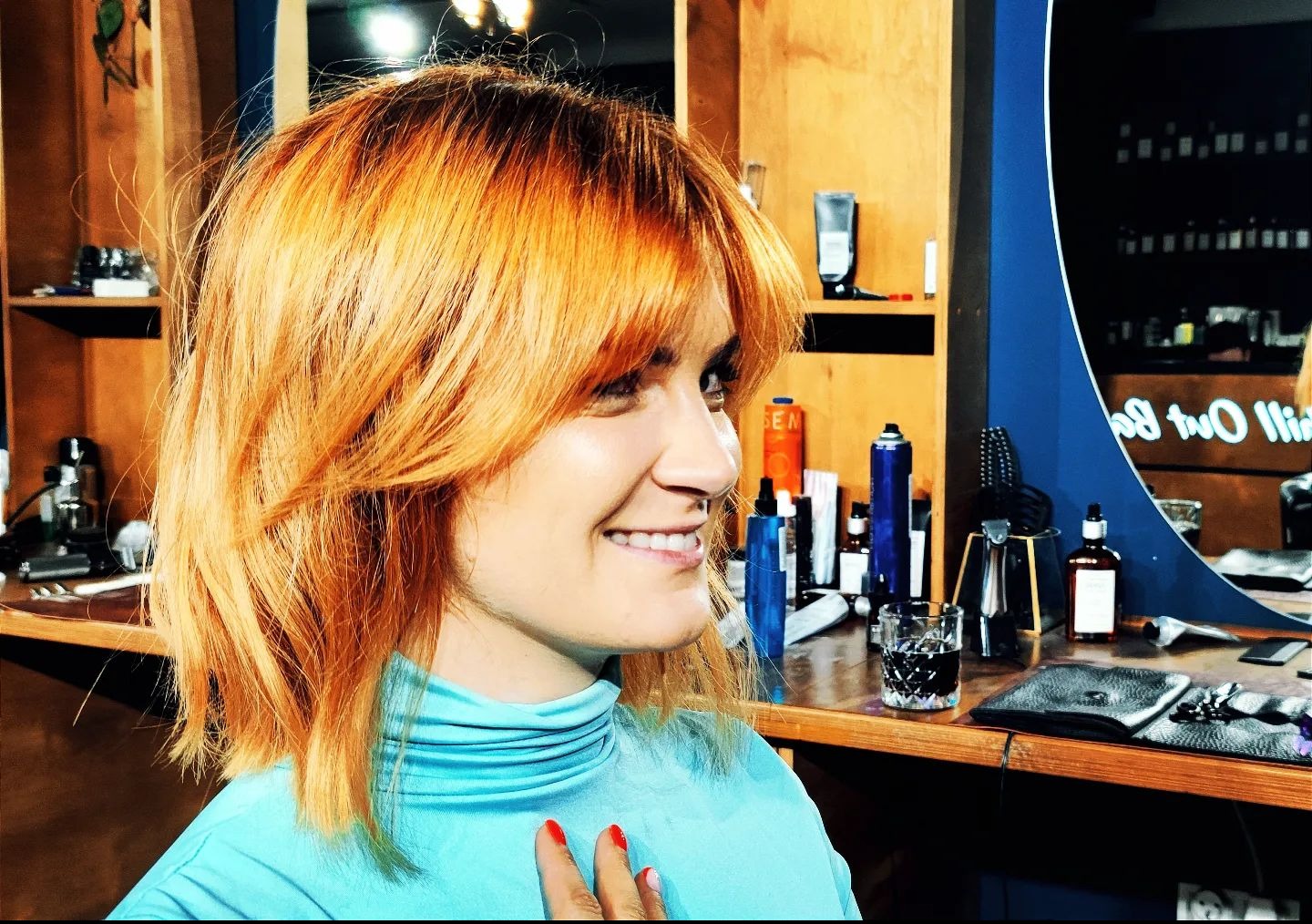 Uśmiechnięta kobieta z rudymi włosami po strzyżeniu, widok z profilu w salonie. Na blacie kosmetyki i akcesoria fryzjerskie.