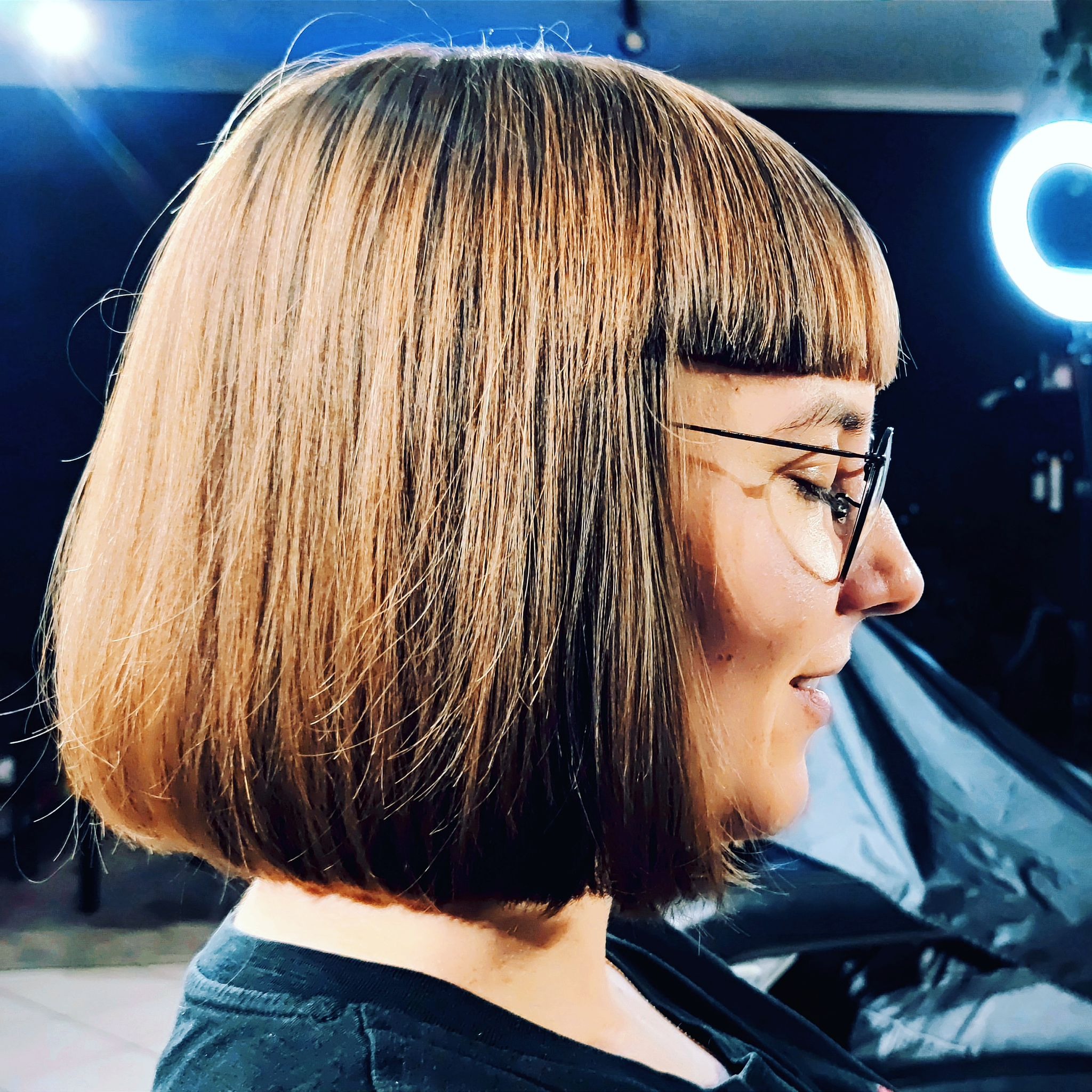 Profil kobiety w okularach z modną, krótką fryzurą w odcieniach blondu i brązu. W tle widoczne oświetlenie studyjne.