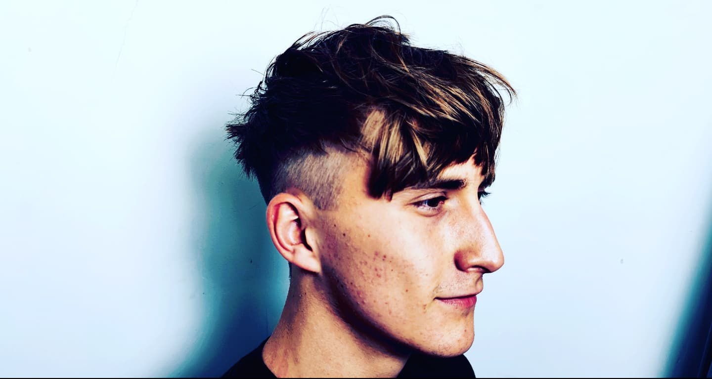 Profil młodego mężczyzny z modną fryzurą: krótko wygolone boki i dłuższa, teksturowana góra z asymetryczną grzywką. Świeże cięcie, widoczne detale.