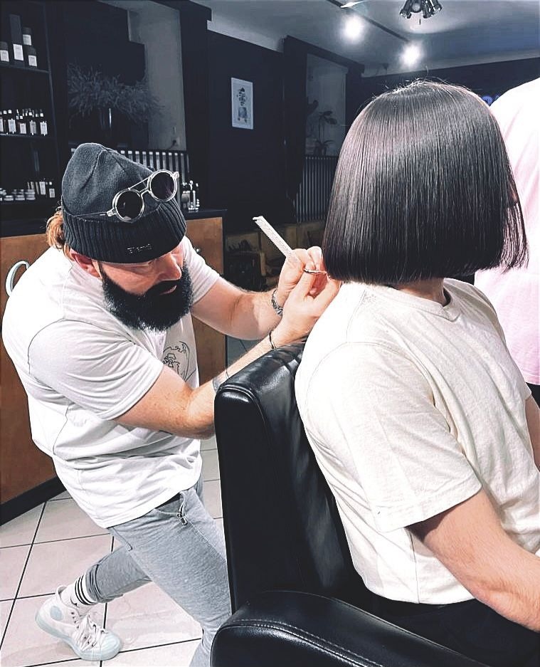 Brodaty mężczyzna w czapce i okularach, strzyże ciemne włosy klientki w salonie. W tle półki z kosmetykami i czarne ściany. Ujęcie z boku, widać detale fryzury.