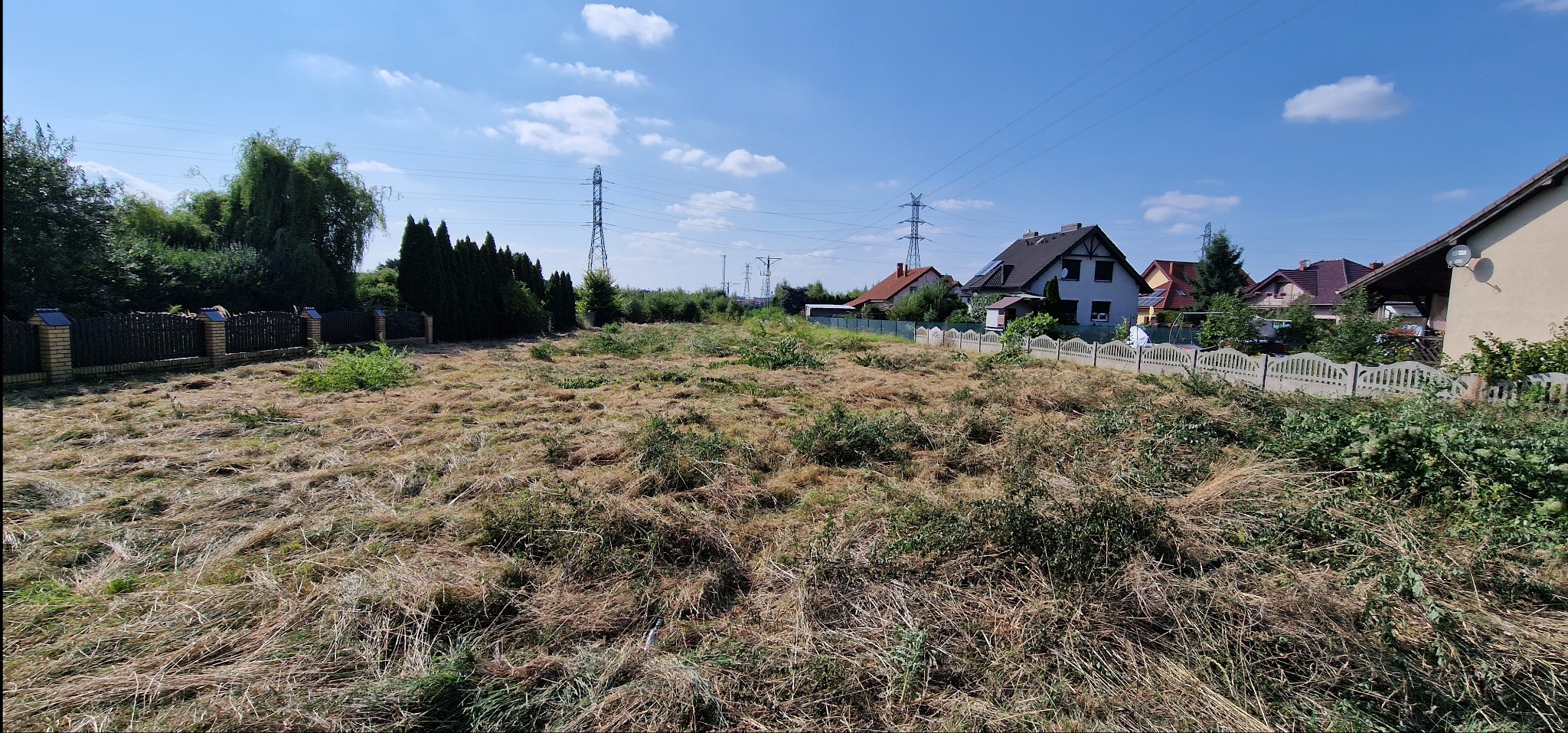 Świeżo skoszone pole z widocznymi śladami trawy, otoczone ogrodzeniami i domami w tle, pod błękitnym niebem z delikatnymi obłokami. Widoczne słupy wysokiego napięcia.