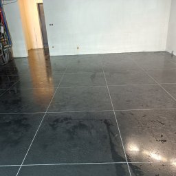Betonvloeren - Błyszcząca, ciemna posadzka z płytek w nowym wnętrzu. Widoczne instalacje grzewcze przy ścianie i świeżo położone płytki z fugami. Minimalistyczny design.