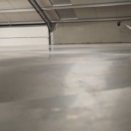 Betonvloeren - Błyszcząca, szara posadzka przemysłowa w hali z otwartą bramą garażową. Na posadzce widoczna maszyna do polerowania, odbijająca się w gładkiej powierzchni.