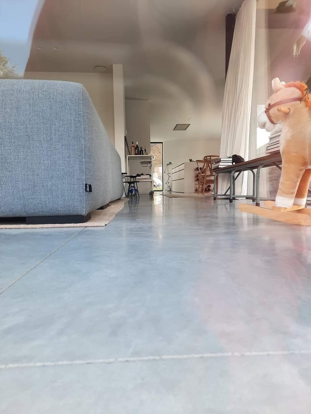 Jasna, gładka posadzka w nowoczesnym wnętrzu z kanapą i zabawką konikiem na biegunach. Minimalistyczny design, widok z niskiej perspektywy podkreśla przestrzeń.