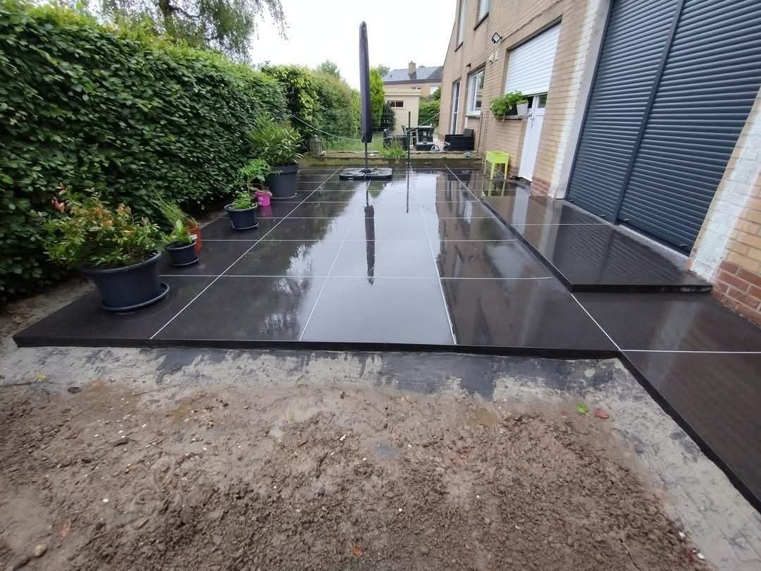 Nowoczesny taras z ciemnych płyt betonowych, z widocznymi fugami i roślinami w donicach. Częściowo wykończony teren wokół tarasu, widoczny fragment budynku.