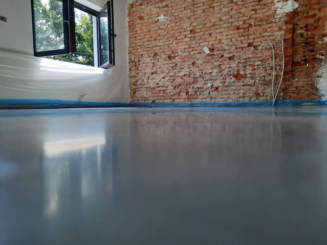 Błyszcząca, szara posadzka w surowym wnętrzu z odsłoniętą cegłą i oknem. Podłoga zabezpieczona folią malarską przy ścianach. Widoczne przewody elektryczne.