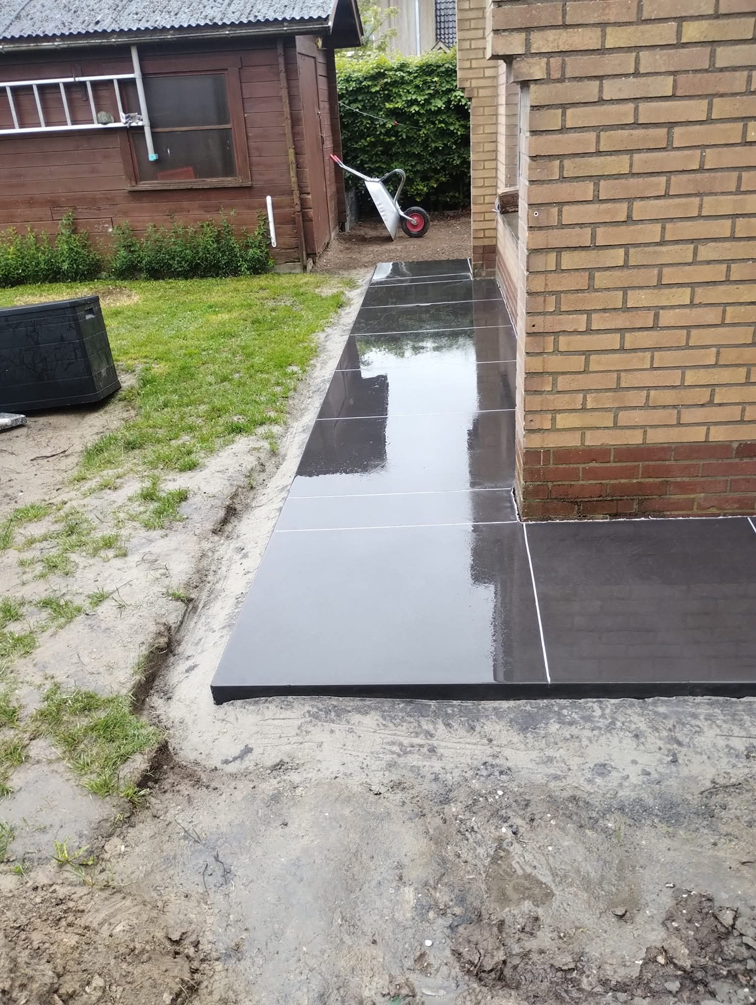 Nowoczesny taras z ciemnych płyt betonowych, z widocznymi fugami, przylegający do ceglanego budynku. W tle zielony ogród i drewniana szopa.