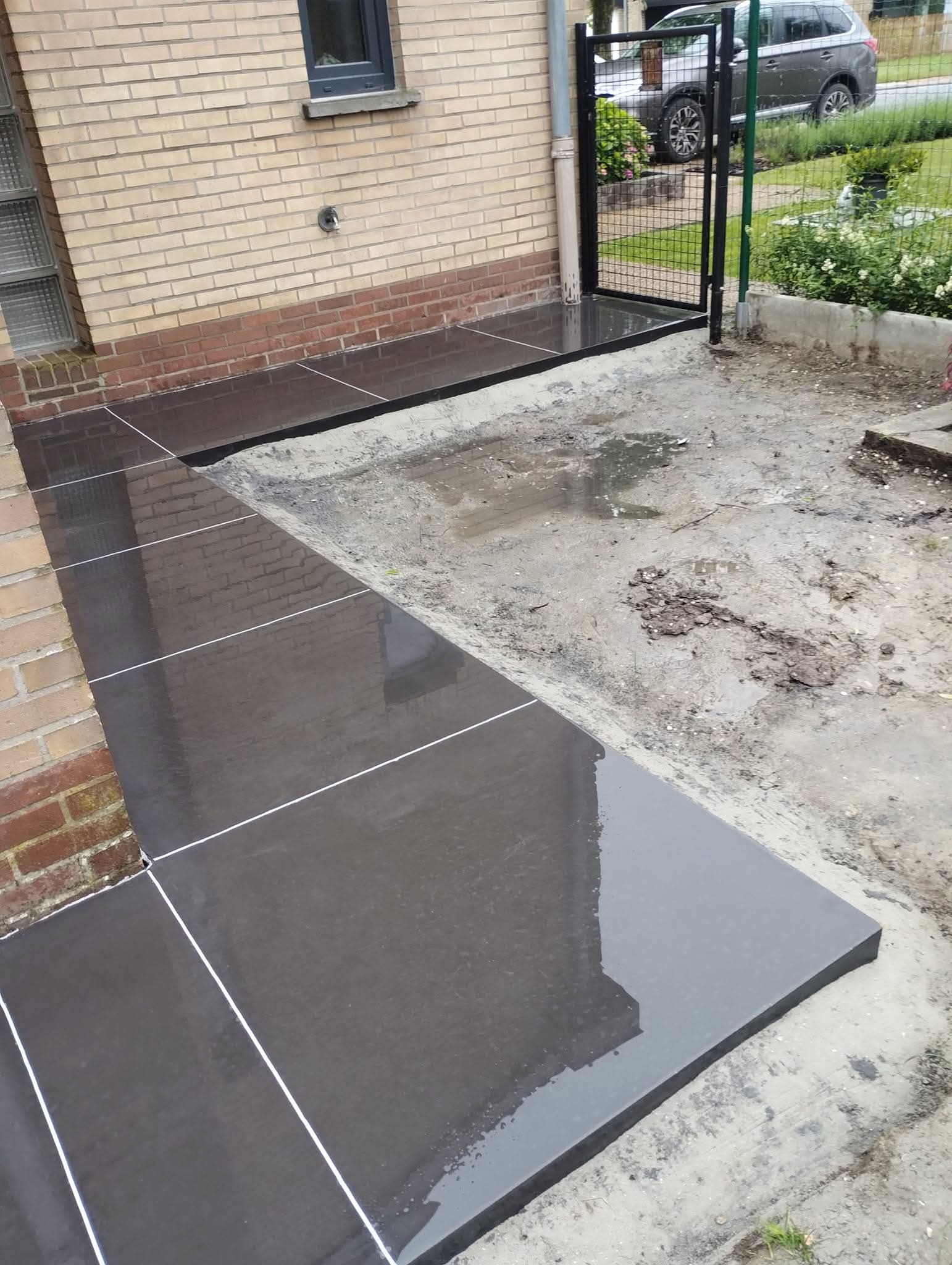 Świeżo ułożony, mokry taras betonowy z ciemnych płyt, z białymi fugami, przylegający do ceglanego budynku. Widoczny fragment ogrodu i ogrodzenia.