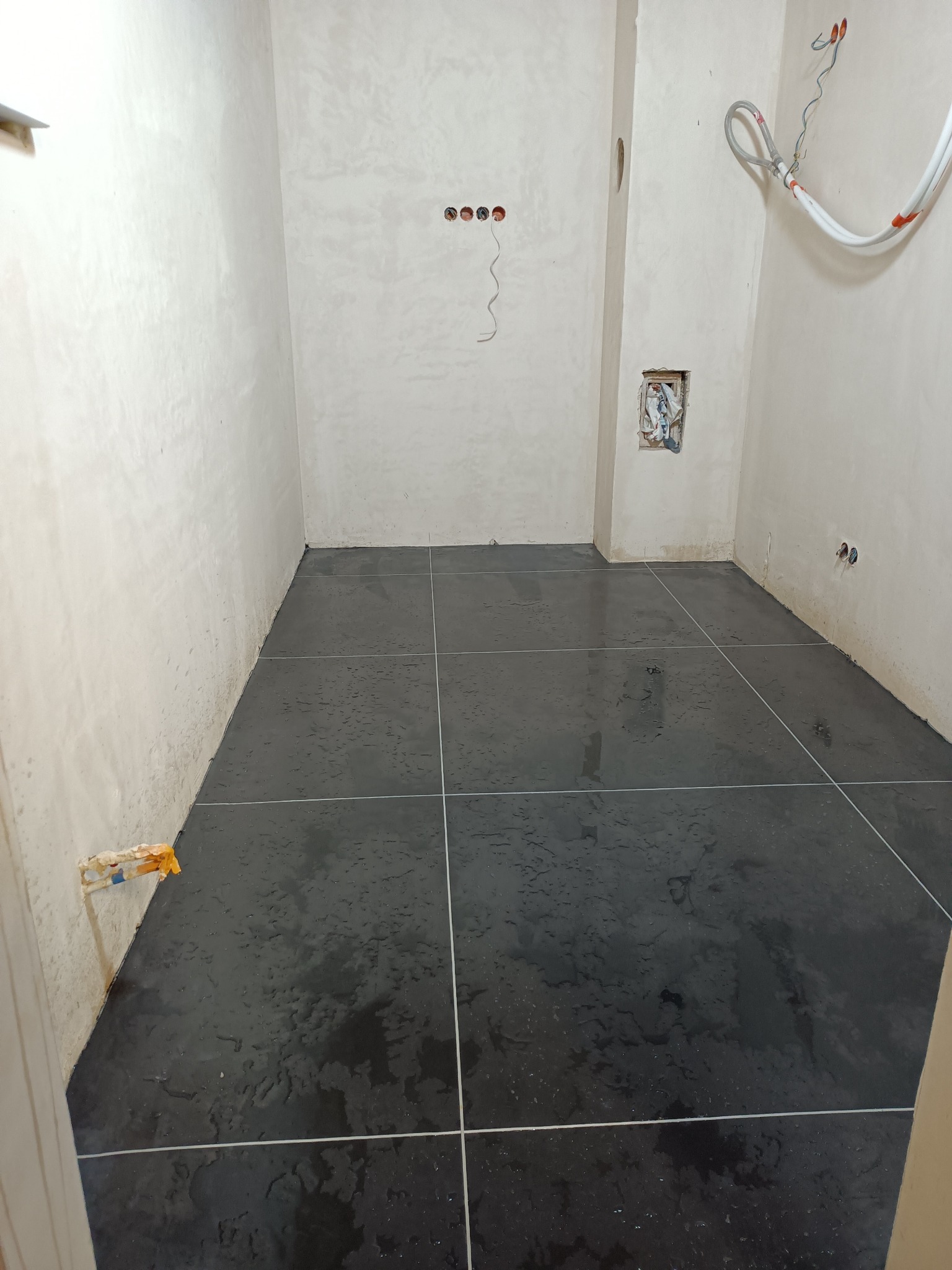Świeżo wylana, ciemna wylewka podłogowa z widocznymi liniami podziału na kwadraty, mokra powierzchnia odbija światło. Ściany surowe, przygotowane do dalszych prac wykończeniowych.
