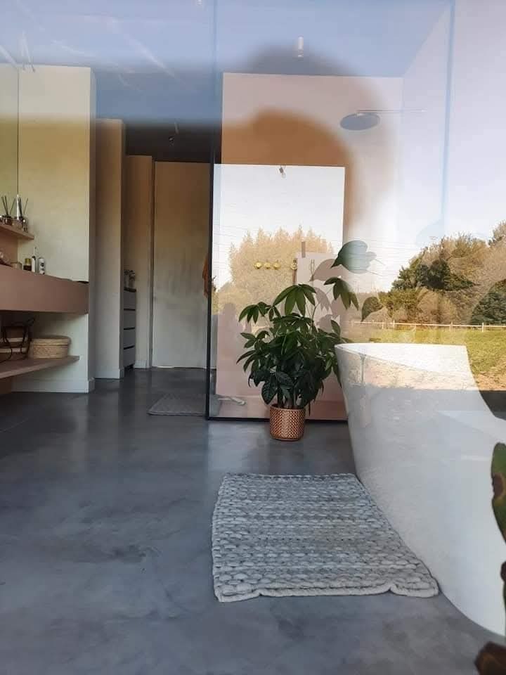 Szara, gładka posadzka w łazience z wanną i rośliną w doniczce. Widoczne odbicie otoczenia w szklanej ścianie. Minimalistyczny design wnętrza.