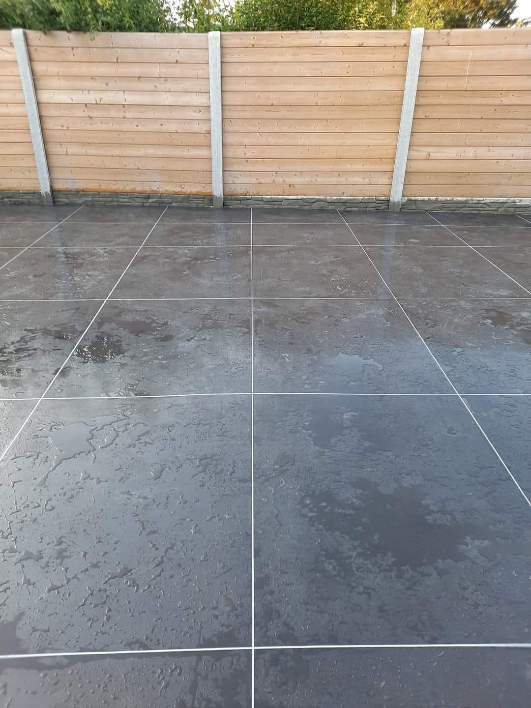 Szare, kwadratowe płyty tarasowe z białymi fugami, mokre od deszczu, przylegające do drewnianego ogrodzenia z betonowymi słupkami. Widok z poziomu podłogi.