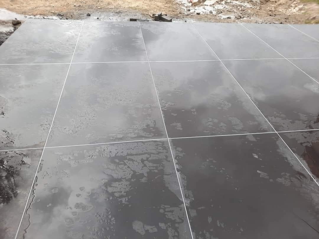 Błyszcząca, ciemna posadzka z wyraźnymi fugami, świeżo po ułożeniu. Widoczne odbicia światła i delikatne nierówności na powierzchni. Tło stanowi ziemia.