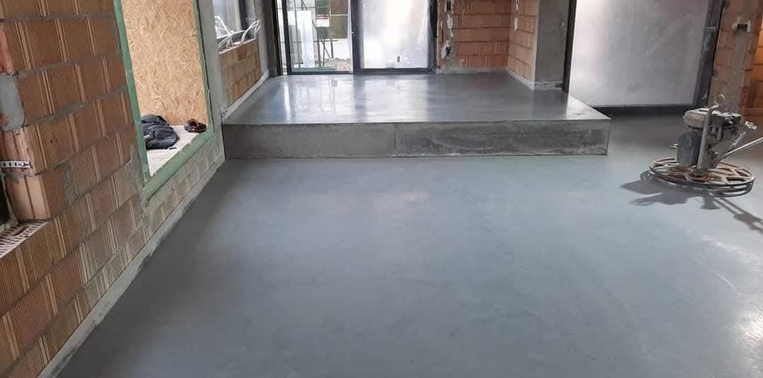 Wypoziomowana, szara wylewka w surowym wnętrzu z ceglanymi ścianami. Na podwyższeniu widoczne drzwi balkonowe. Na pierwszym planie maszyna do polerowania.