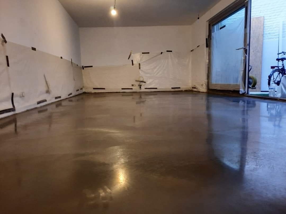 Świeżo wylana, błyszcząca posadzka w pomieszczeniu z zabezpieczonymi ścianami folią i taśmą. Widoczne odbicia światła na gładkiej powierzchni.