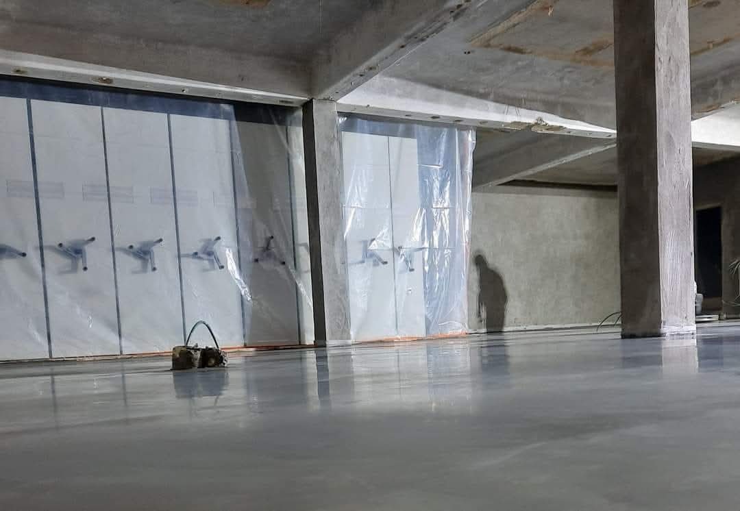 Gładka, szara powierzchnia w hali z kolumną i osłoniętymi folią ścianami. Widoczny cień osoby i sprzęt do obróbki. Świeżo wykonana wylewka.