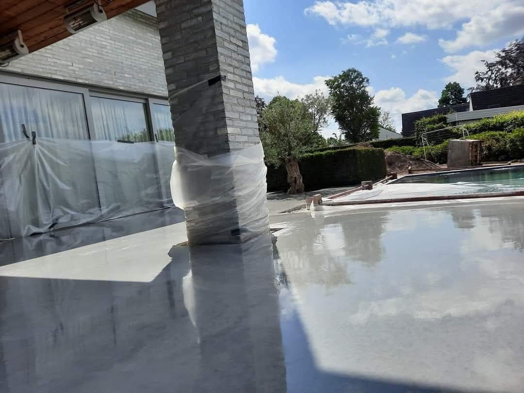 Świeża, szara wylewka na tarasie zewnętrznym, z kolumną zabezpieczoną folią. W tle basen i zieleń, widoczne odbicie nieba w mokrej powierzchni.