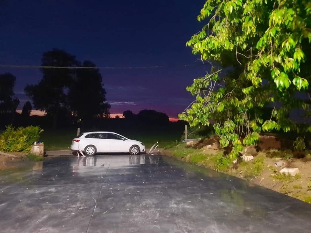 Świeżo wylany, mokry taras betonowy o gładkiej powierzchni, odbijający nocne niebo. Obok stoi biały samochód, a w tle widać zieleń drzew i krzewów.