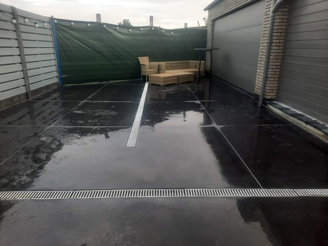 Gładka, ciemna powierzchnia z betonu podzielona na kwadraty, z widocznymi odpływami liniowymi. W tle drewniane meble ogrodowe i brama garażowa.