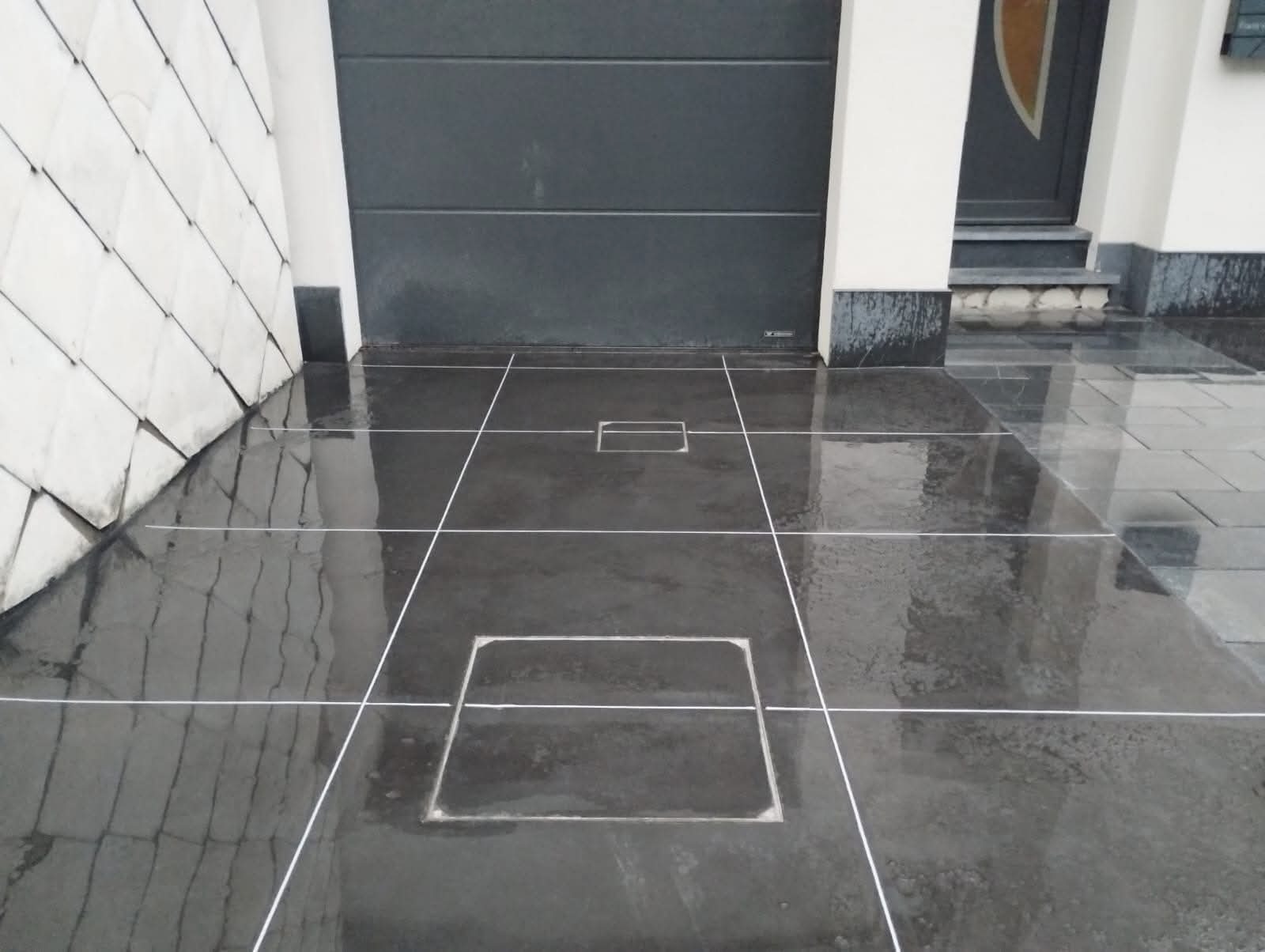 Błyszcząca, ciemna powierzchnia z kwadratowymi wnękami i białymi liniami podziału, odbijająca otoczenie. Widoczny fragment ściany z szarej cegły i brama garażowa.