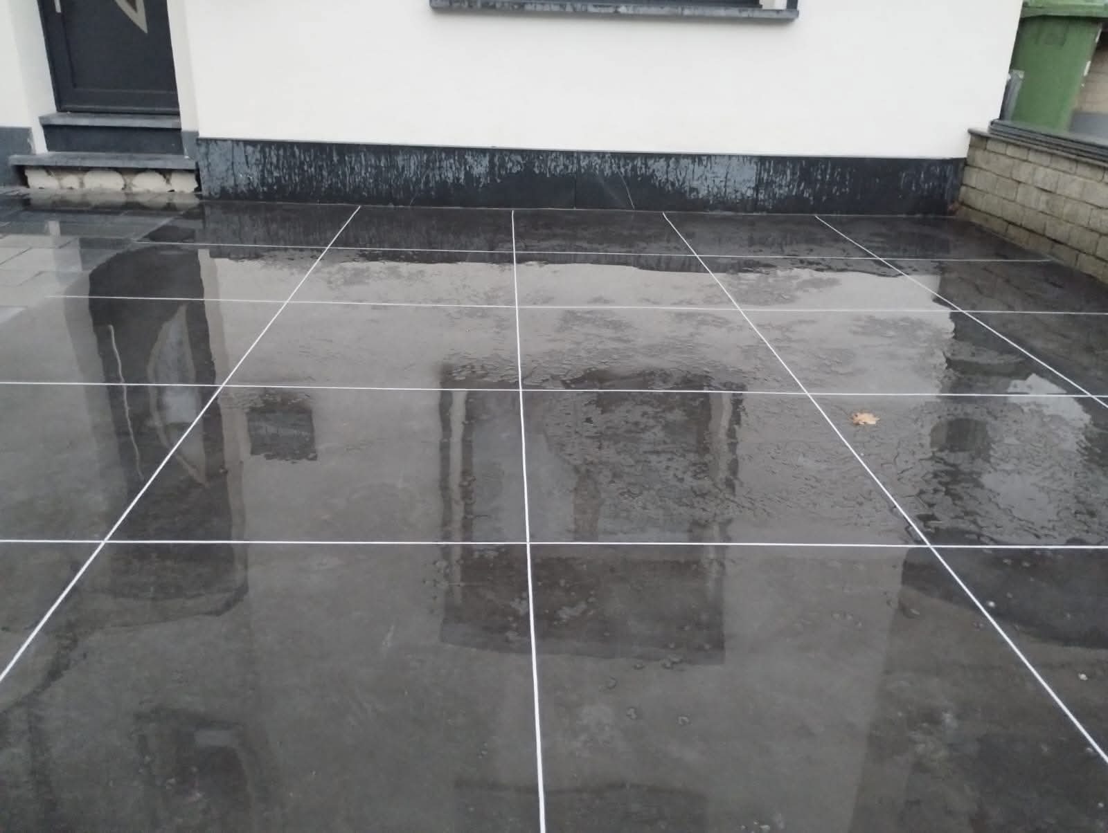 Ciemnoszara, mokra nawierzchnia z płyt, z widocznymi białymi liniami fug, odbijająca otoczenie. Widoczny fragment ściany budynku i drzwi wejściowe.