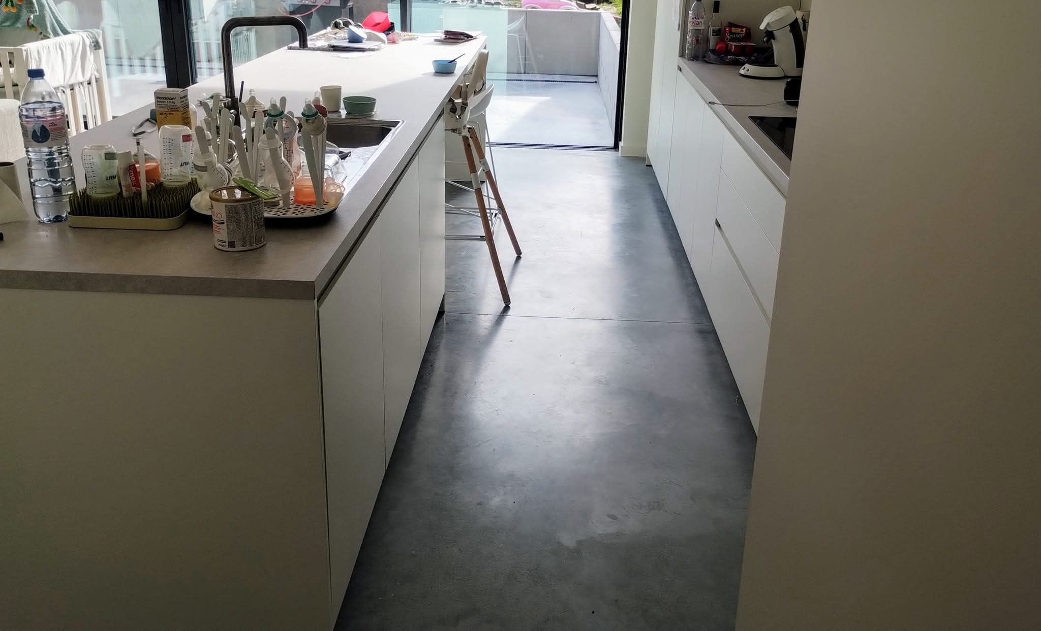 Szara, gładka podłoga w nowoczesnej kuchni z białymi szafkami i wyspą. Widoczne akcesoria kuchenne i krzesełko dziecięce. Minimalistyczny design, jasne wnętrze.