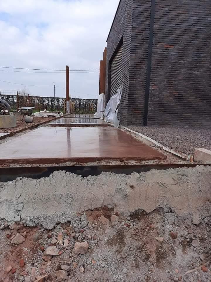 Wylewka betonowa na zewnątrz budynku z cegły, widoczne przygotowania do dalszych prac. Częściowo zakryte folią elementy. Ujęcie z bliska, ziemia i gruz w kadrze.