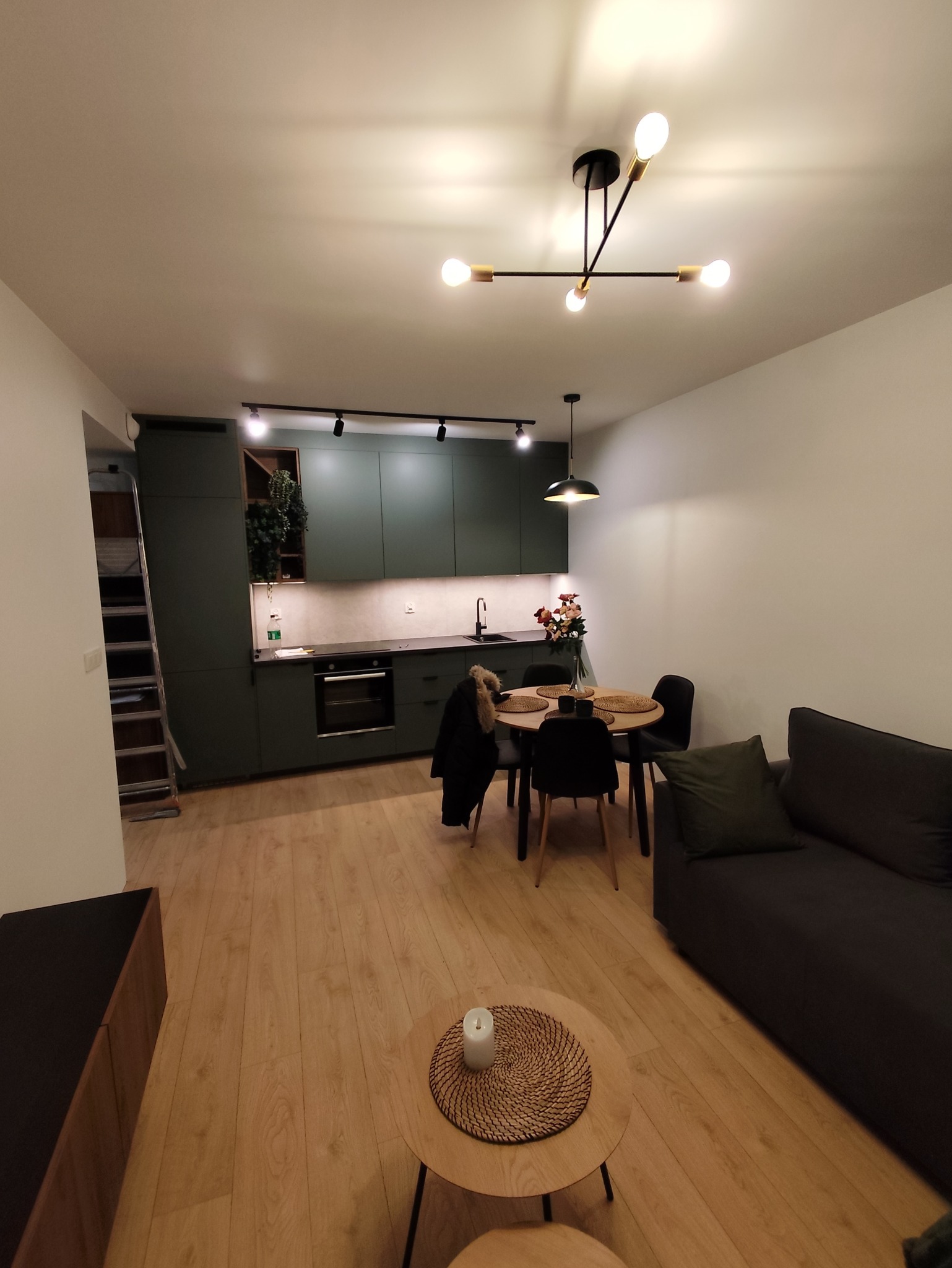 Nowoczesne wnętrze po remoncie: zielona zabudowa kuchenna, drewniana podłoga, stół z krzesłami i sofa. Minimalistyczna lampa sufitowa dodaje elegancji.