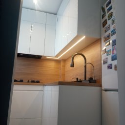 Tomek Banasiak - Nowoczesna kuchnia z białymi szafkami i drewnianym blatem, oświetlona listwą LED. Na ścianie lodówka z magnesami. Widok zza framugi.