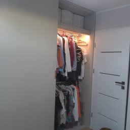 Tomek Banasiak - Otwarta szafa wnękowa z przesuwnymi drzwiami, oświetleniem wewnętrznym i ubraniami na wieszakach. Obok widoczny fragment drzwi i leżący pies.