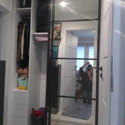 Tomek Banasiak - Biała szafa wnękowa z czarnymi, przesuwnymi drzwiami z lustrem, w którym odbija się osoba robiąca zdjęcie. Widoczne półki z ubraniami i akcesoriami.