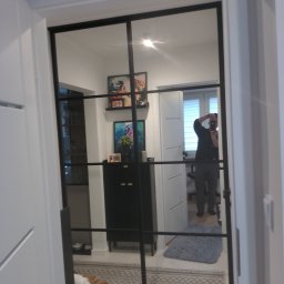 Tomek Banasiak - Nowoczesne drzwi przesuwne z czarnymi szprosami, odbijające wnętrze pokoju z szafką, obrazami i osobą robiącą zdjęcie. Widoczny fragment dywanu i drzwi.