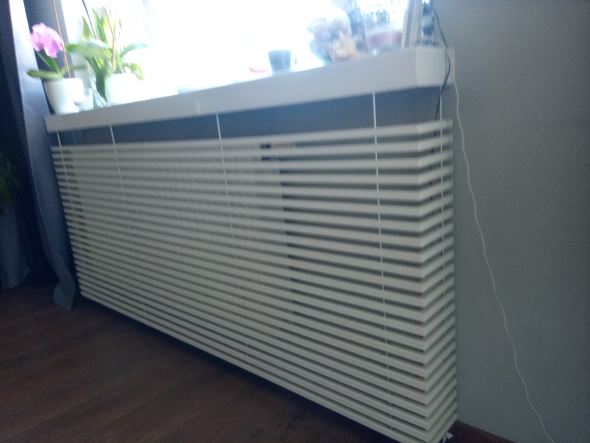 Biały, poziomy grzejnik z żaluzjami w pokoju, z kwiatami na parapecie. Widoczne drewniane panele podłogowe i szara ściana. Kabel elektryczny zwisa po prawej stronie.