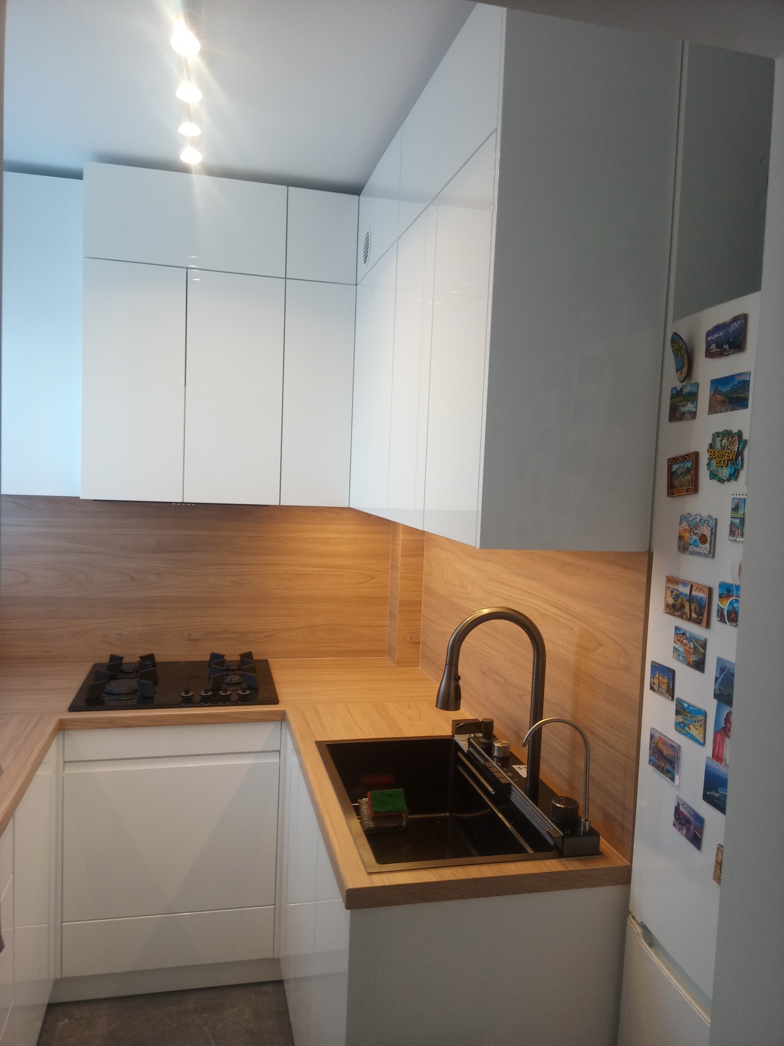 Nowoczesna kuchnia z białymi szafkami, drewnianym blatem i czarnym zlewem. Nad blatem rząd białych szafek wiszących. Na lodówce magnesy z podróży.