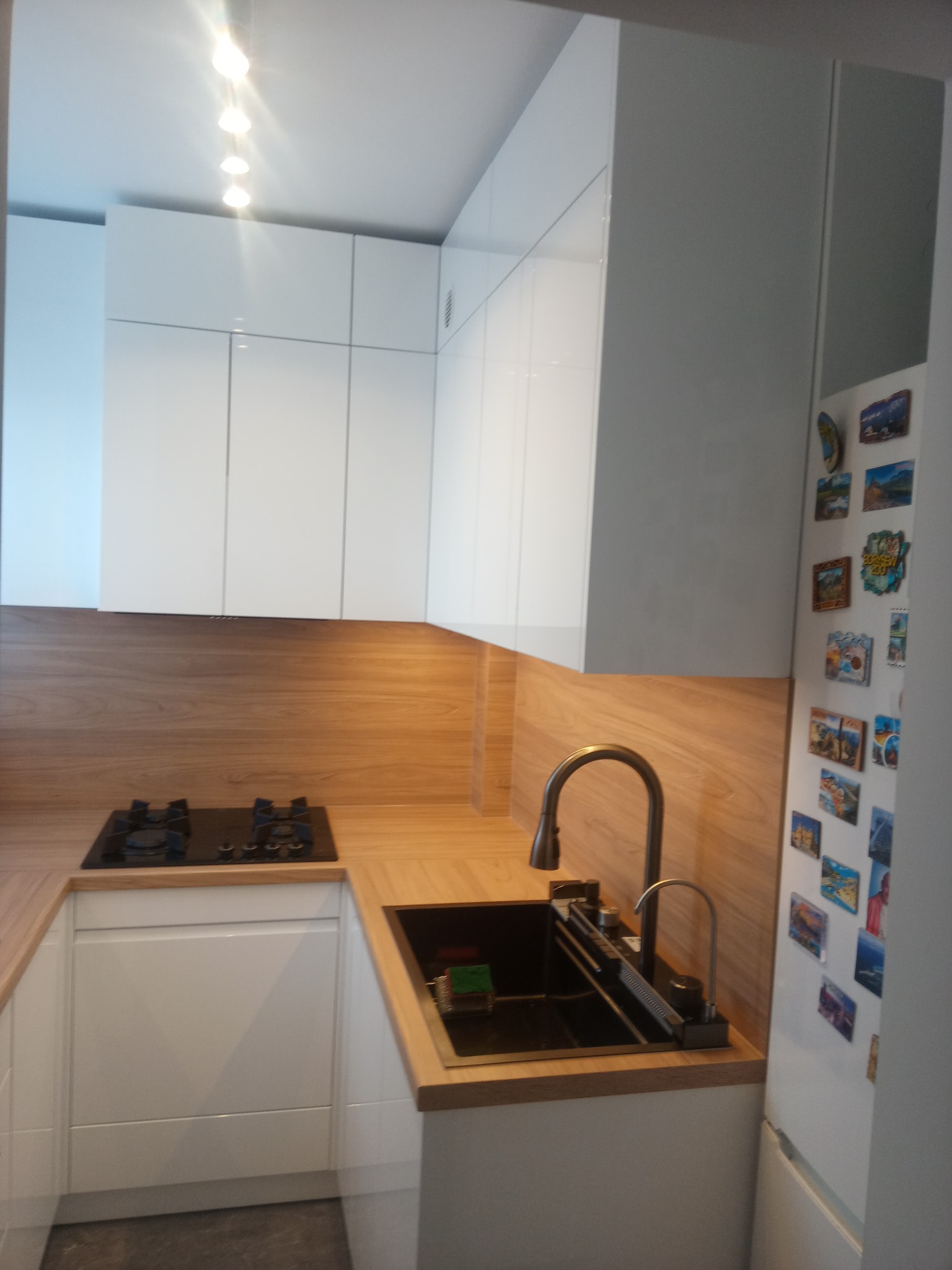 Nowoczesna, jasna kuchnia z białymi szafkami i drewnianym blatem. Widoczny zlew z baterią, płyta gazowa i lodówka z magnesami. Minimalistyczny design.