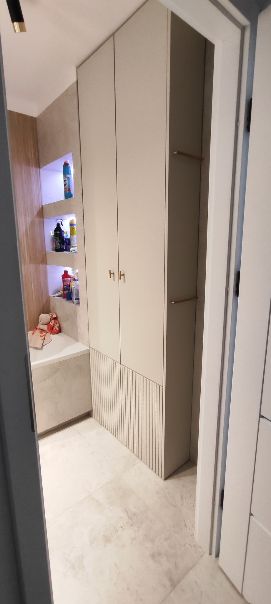 Beżowa szafa wnękowa z ryflowanym dołem w łazience z półkami wnękowymi. Widok zza uchylonych drzwi.