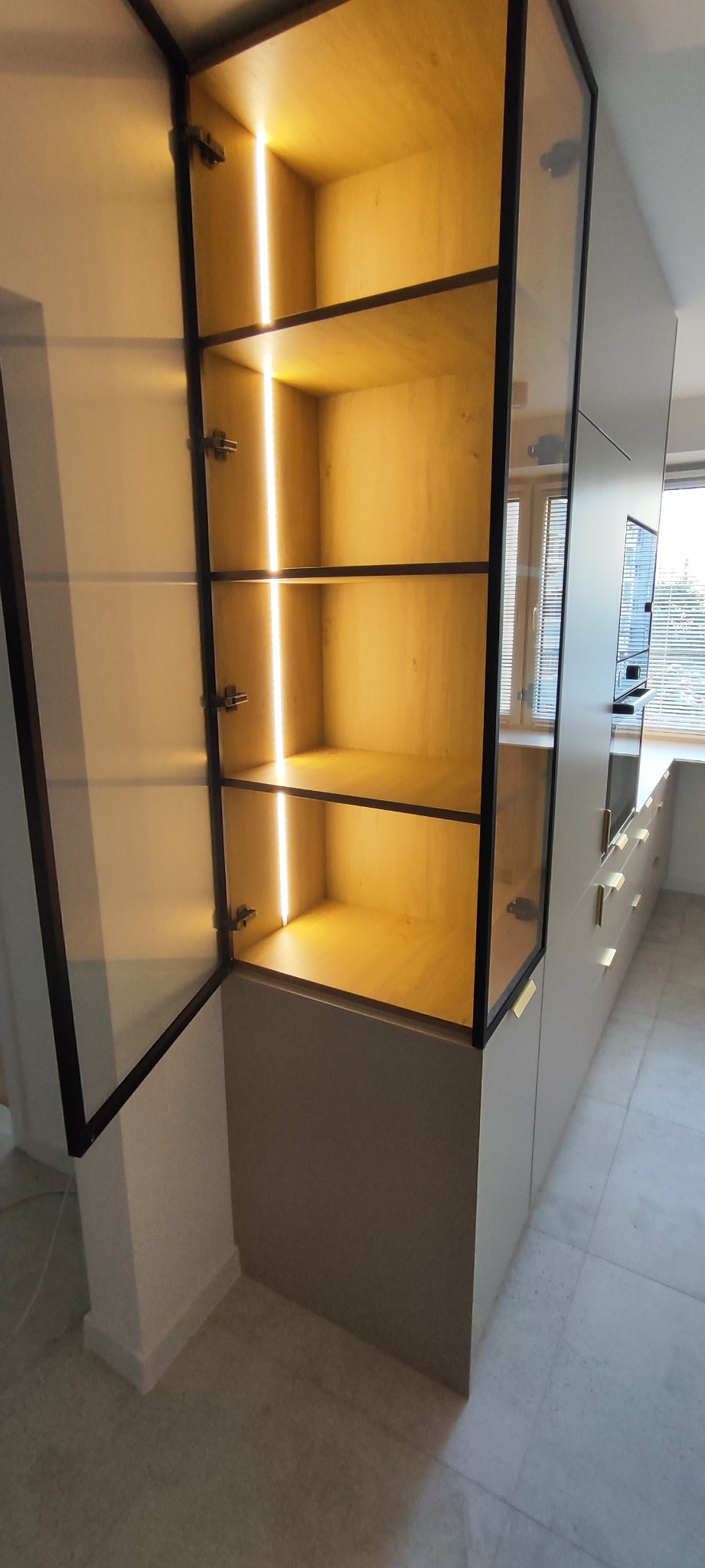 Otwarta szafka kuchenna z podświetlanymi półkami, czarne obramowanie szklanych drzwi, beżowe fronty szafek, widok na okno w tle.