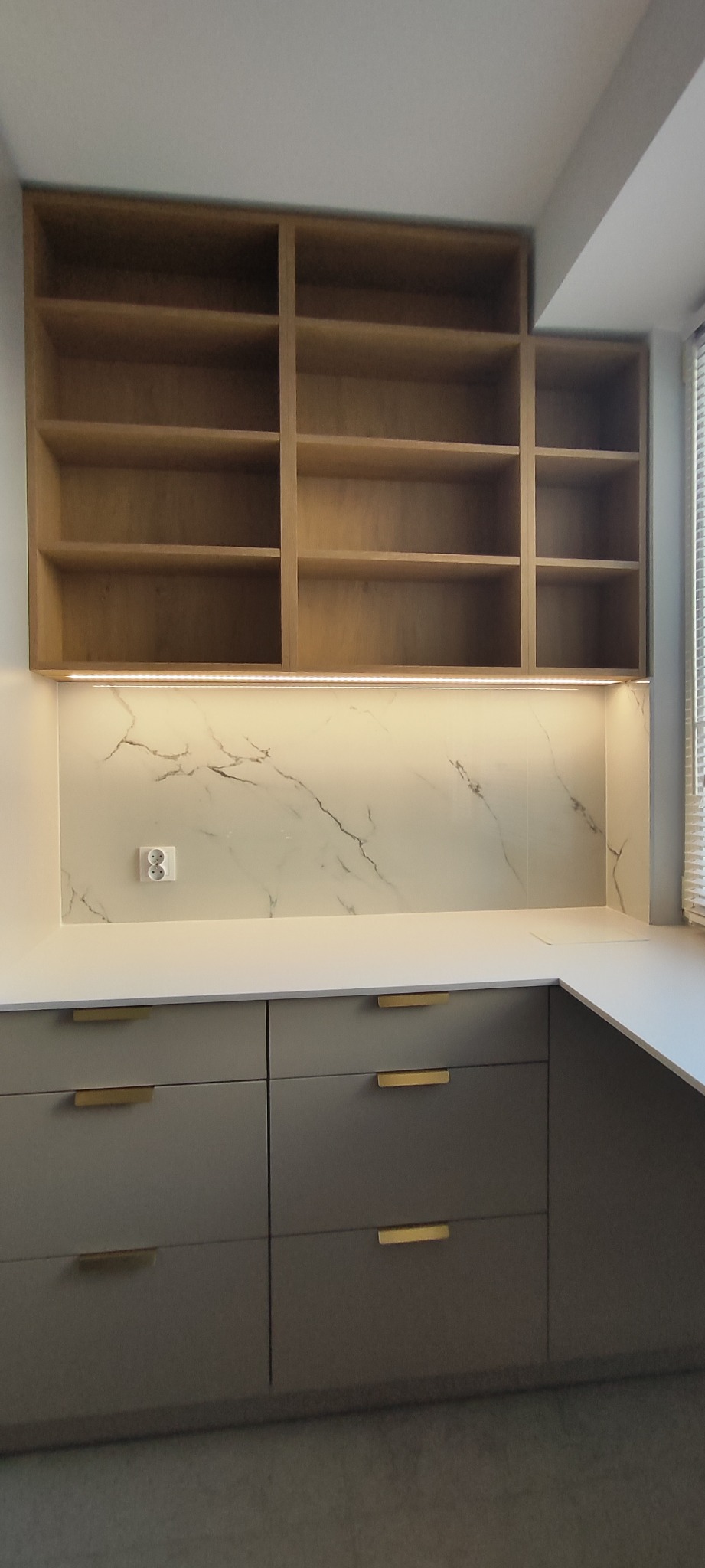 Szare szafki kuchenne z złotymi uchwytami, biały blat i marmurowy panel nad blatem, oświetlenie LED pod wiszącą, drewnianą półką z otwartymi przegródkami.