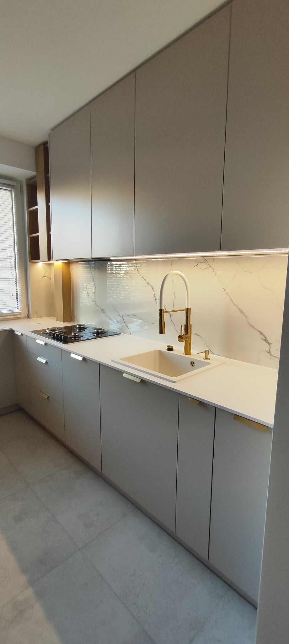 Jasna kuchnia z szarymi szafkami, złotą armaturą i blatem imitującym marmur. Widoczne oświetlenie podszafkowe i płyta grzewcza. Minimalistyczny design.