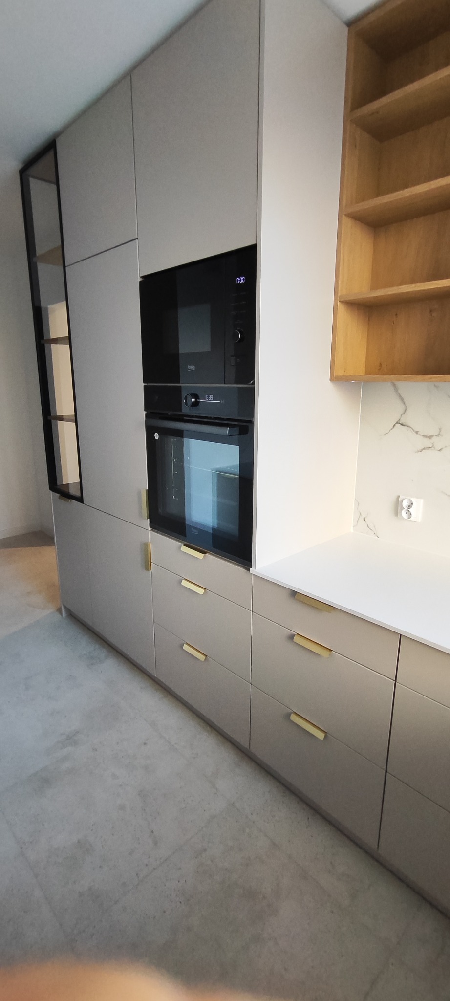 Jasna zabudowa kuchenna z piekarnikiem i mikrofalówką w czarnych obudowach. Szafki z podłużnymi, złotymi uchwytami. Otwarta szafka z drewnianymi półkami po prawej.